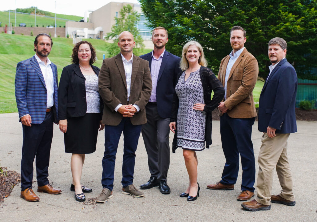 The Agile5 Team Group photo of the seven Agile5 team members. The group is standing outdoors and the image shows a diverse mix of men and women in business attire, standing in a line on a path with grass and trees in the background.